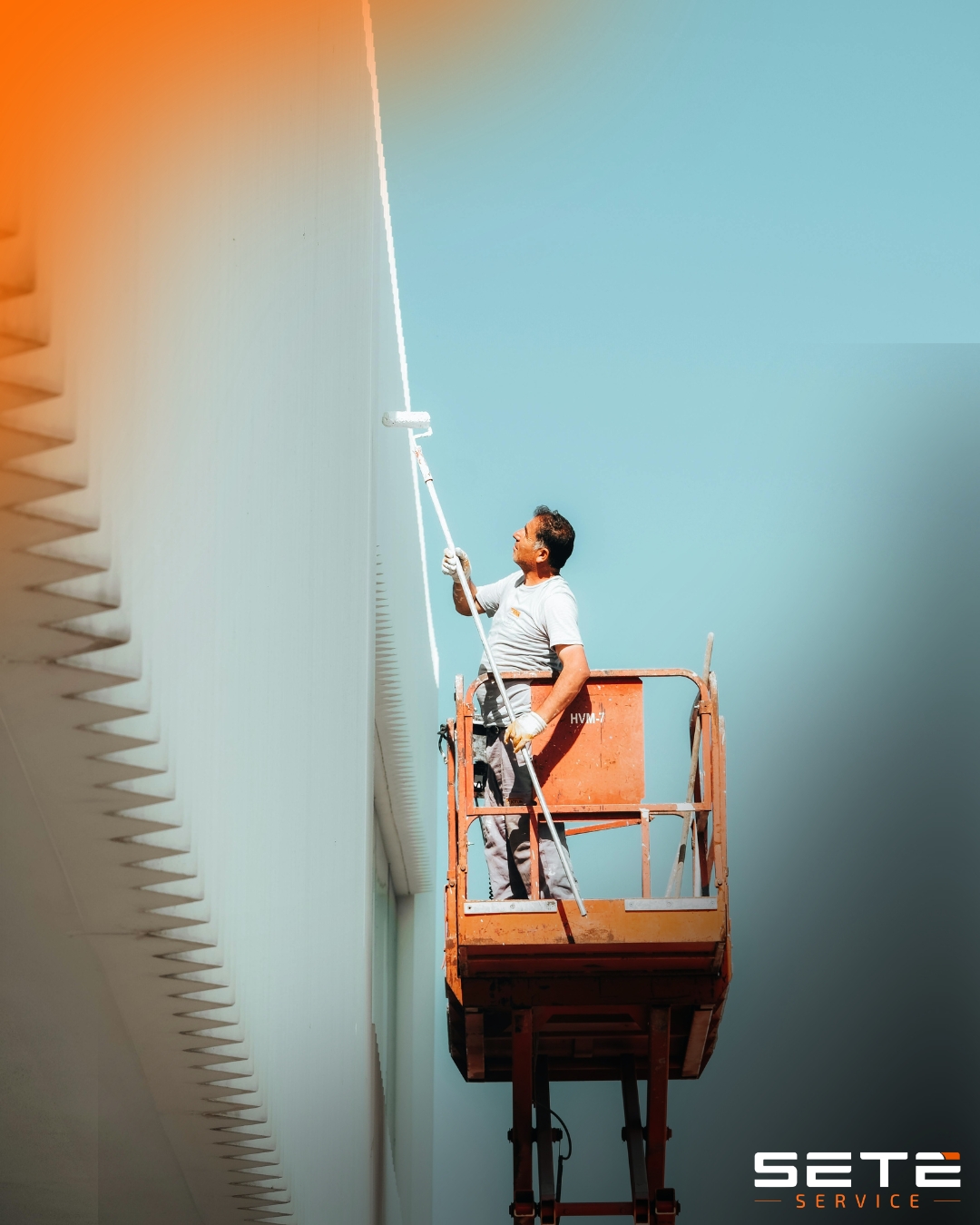 Pintor em plataforma de trabalho aéreo aplica tinta branca em parede alta contra céu azul claro.